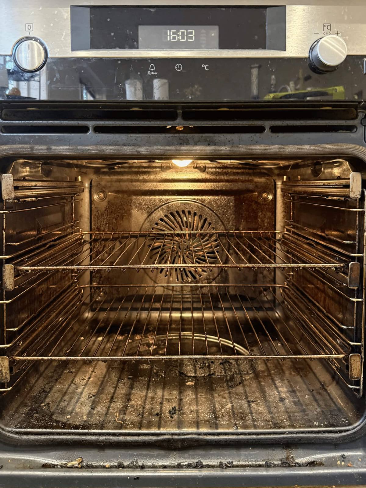 Wall oven at 16:03 with the door open, racks in place and the cavity coated in baked-on grease