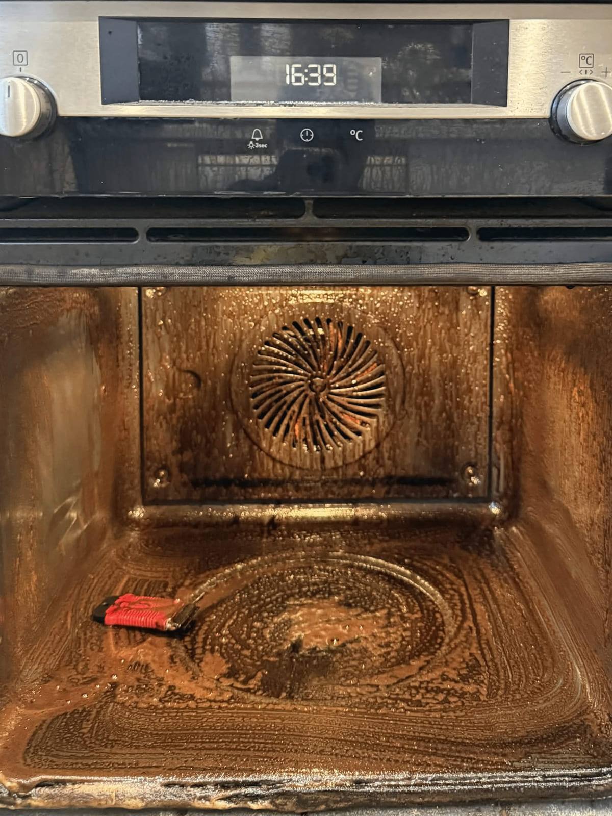 The oven cavity at 16:39 with softened grease lifting away as it is scraped clear by hand