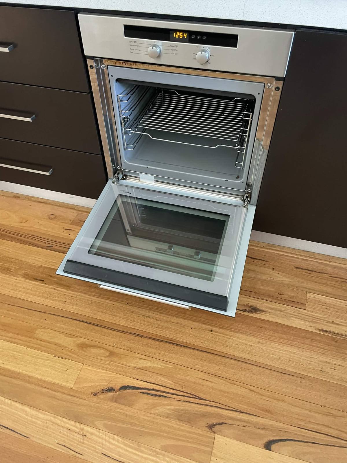 Stainless steel wall oven with the door open after cleaning, clean racks and clear glass, over timber flooring