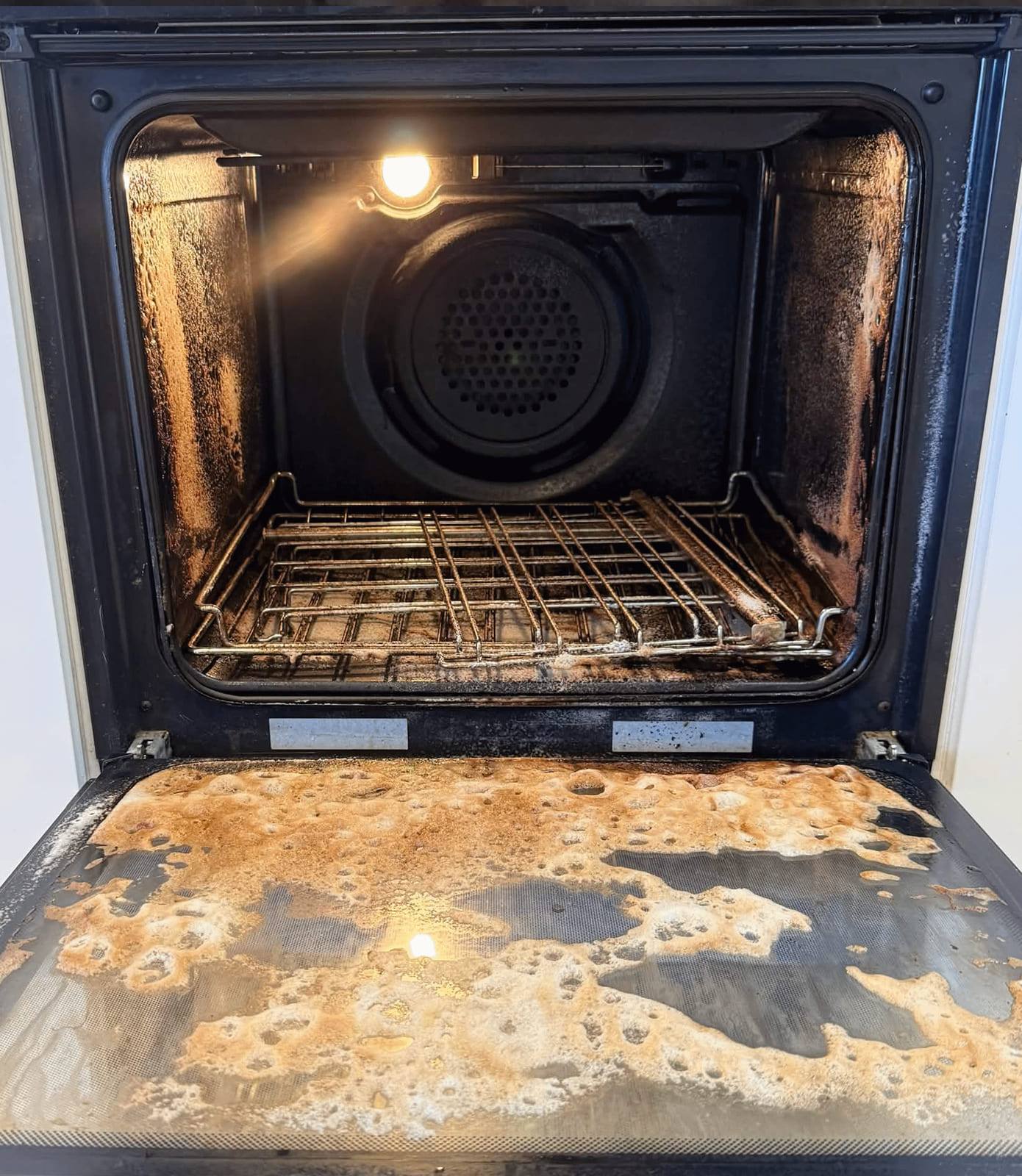 Lit wall oven cavity with chrome racks above a door tray covered in orange baked-on spills